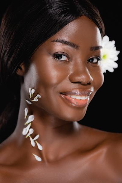nahá africká americká žena s bílou chryzantémou ve vlasech a okvětními lístky na krku izolované na černé - Fotografie, Obrázek
