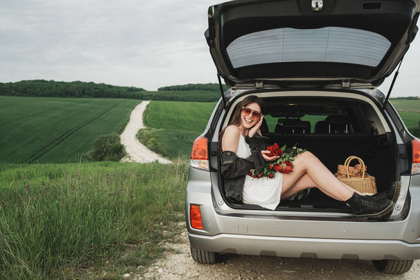 Mladá stylová žena v bílých šatech a džínové bundy s kyticí červených pivoňky sedí na kufru svého SUV auta v zeleném poli - Fotografie, Obrázek