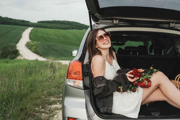 Joven mujer con estilo en vestido blanco y chaqueta de mezclilla con ramo de peonías rojas se sienta en el maletero de su coche SUV en el campo verde - Foto, Imagen
