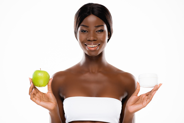 mujer afroamericana sosteniendo manzana verde madura y crema cosmética aislada en blanco - Foto, imagen