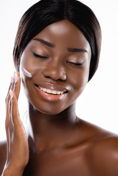 Αφροαμερικανή γυμνή γυναίκα με κλειστά μάτια που απλώνει καλλυντική κρέμα στο πρόσωπο που απομονώνεται σε λευκό - Φωτογραφία, εικόνα