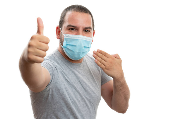Cheerful male adult presenting disposable surgical or medical face mask making thumb up like gesture with hand as covid19 influenza pandemic concept isolated on white background - 写真・画像