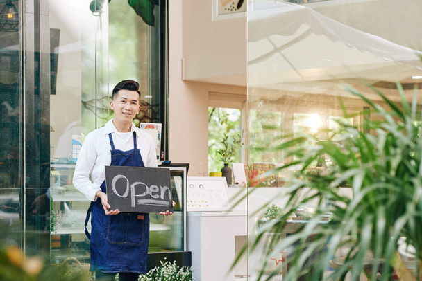 Sonriente camarero feliz café de pie en la entrada y mostrando signo abierto al dar la bienvenida a los clientes después del bloqueo ha terminado - Foto, Imagen