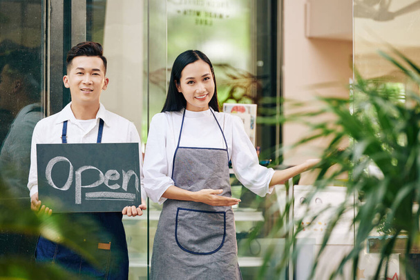 Улыбающийся молодой официант показывает открытый знак, когда официантка делает приветственный жест и приглашает клиентов внутрь - Фото, изображение