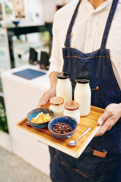 Обрізане зображення офіціанта, що приносить лоток зі сніданком, що складається з йогурту, кукурудзяних пластівців та шоколадних чіпсів
 - Фото, зображення