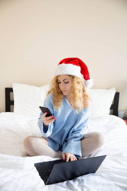 Žena v Santa Claus červená čepice s chytrým telefonem a notebookem v posteli. - Fotografie, Obrázek