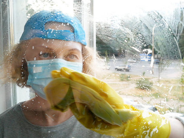 Γυναίκα με προστατευτική μάσκα και γάντια πλύσιμο παραθύρων. Έννοια υπηρεσιών καθαρισμού παραθύρων. - Φωτογραφία, εικόνα