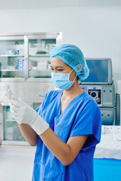 Ernstige jonge chirurgische assistent in scrubs en medisch masker zetten gesteriliseerde chirurgische handschoenen operatiekamer - Foto, afbeelding