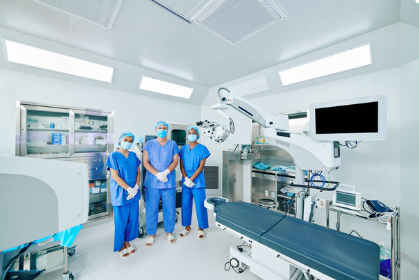 Vážní zdravotníci v chirurgické uniformě, maskách a rukavicích stojící na operačním sále - Fotografie, Obrázek