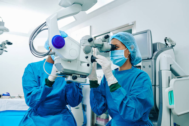 Ασιάτισσα χειρουργός με τρίψιμο που χρησιμοποιεί ηλεκτρονικό μικροσκόπιο όταν χειρουργεί ασθενή - Φωτογραφία, εικόνα