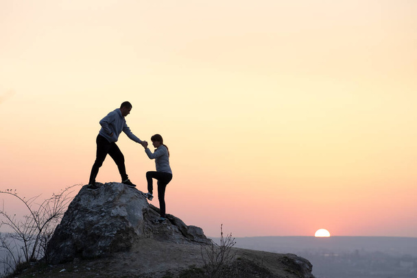 Férfi és női túrázók segítik egymást, hogy sziklát másszanak naplementekor a hegyekben. Egy pár mászik a magas sziklára az esti természetben. Turizmus, utazás és egészséges életmód koncepció. - Fotó, kép