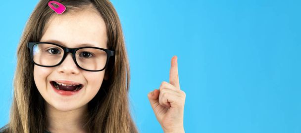 Close up portrait of a child school girl wearing looking glasses holding up point finger in I have an idea gesture isolated on blue background. - Photo, Image