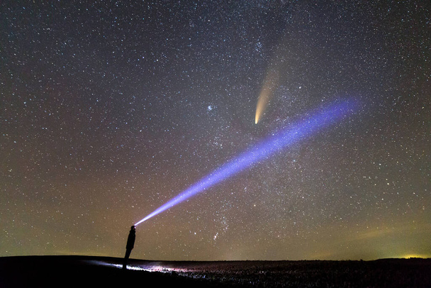 Silhouette eines Mannes mit Taschenlampe auf dem Kopf, die einen hellen Lichtstrahl auf den Sternenhimmel mit dem Kometen C / 2020 F3 (NEOWISE) mit hellem Schweif zeigt. - Foto, Bild