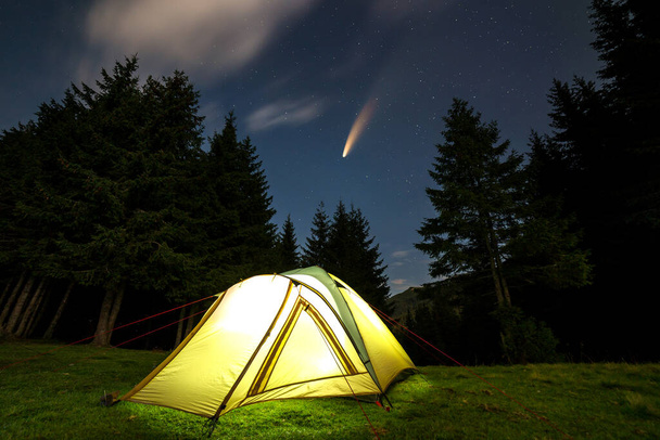 Φωτισμένη τουριστική σκηνή σε πράσινο ξέφωτο με μακρινό βουνό και έναστρο νυχτερινό ουρανό με Neowise κομήτη με ελαφριά ουρά σε αυτό. - Φωτογραφία, εικόνα