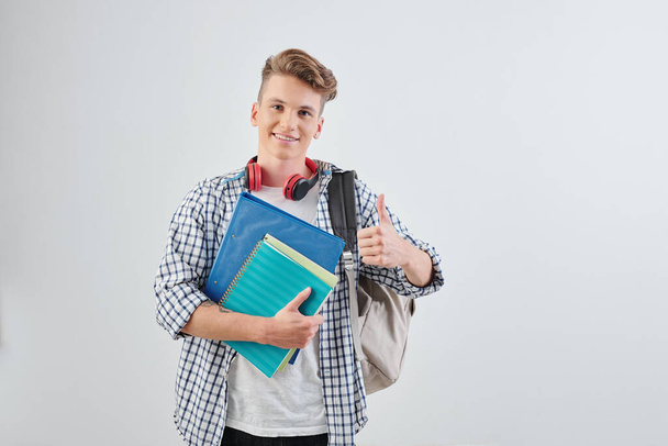 Portrait of happy handsome young high school student with textbooks showing thumbs-up and smiling at camera - Photo, image
