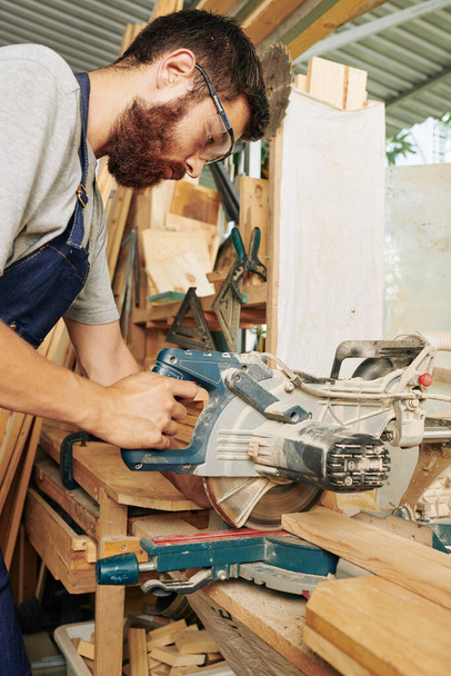 Σοβαρός επαγγελματίας ξυλουργός που χρησιμοποιεί κυκλικό πριόνι σκουληκιών για την κοπή ξύλινων σανίδων για το έργο του - Φωτογραφία, εικόνα