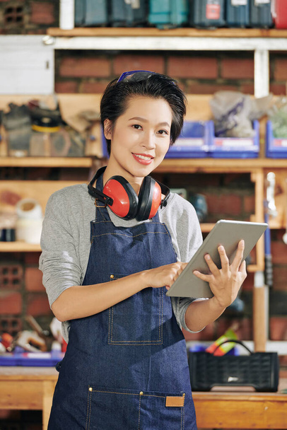 Portrait de sourire jolie jeune charpentier asiatique en tablier de denim en utilisant l'application sur tablette pour vérifier l'ordre des clients - Photo, image