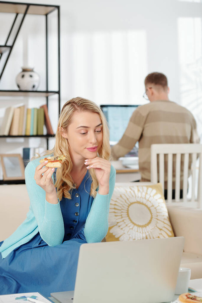 Nuori nainen syö pizzaa ja tarkistaa sähköpostit, kun hänen miehensä työskentelee tietokoneella taustalla - Valokuva, kuva