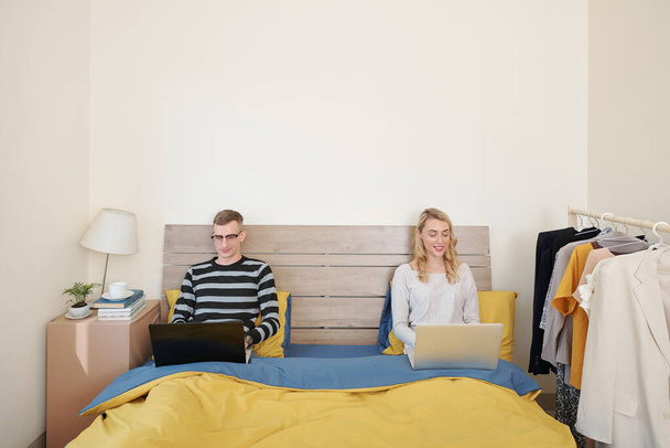 Gelukkig jong man en vrouw zitten op bed met laptops, werken of spelen van games online - Foto, afbeelding