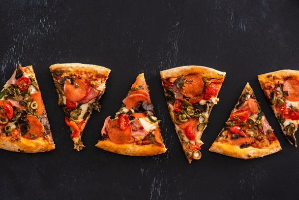 вид сверху на вкусные итальянские ломтики пиццы с овощами и салями на черном фоне - Фото, изображение
