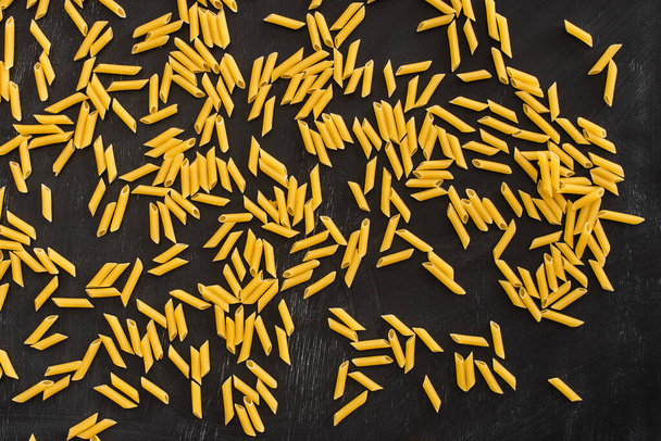 bovenaanzicht van rauwe penne pasta verspreid over zwarte achtergrond - Foto, afbeelding