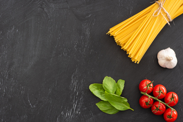top view of raw Italian spaghetti with basil leaves, tomatoes and garlic on black background - Foto, afbeelding