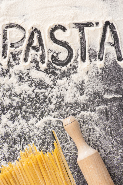bovenaanzicht van rauwe spaghetti bij het rollen pin en pasta woord op bloem op zwarte achtergrond - Foto, afbeelding