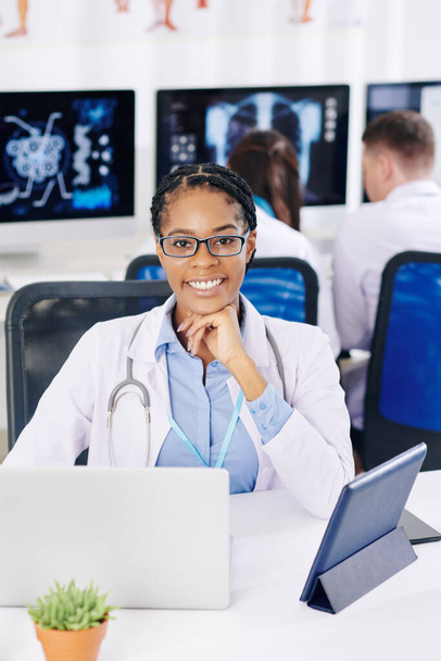 Pozitivní sebevědomá afroamerická praktická lékařka sedící za stolem v laboratoři a pracující na počítači - Fotografie, Obrázek