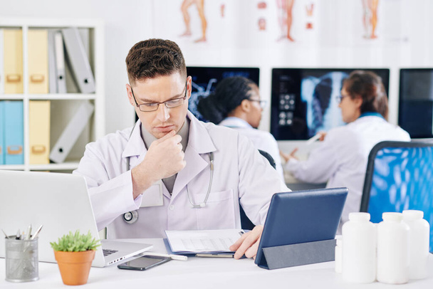 Серьезный хмурый врач проверяет документы со статистикой заболеваемости, сидя за столом - Фото, изображение