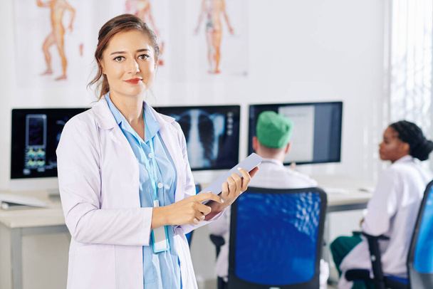 Pozitív fiatal háziorvos portréja, aki táblagépet tart, és orvosi rendelőben állva mosolyog a kamera előtt - Fotó, kép
