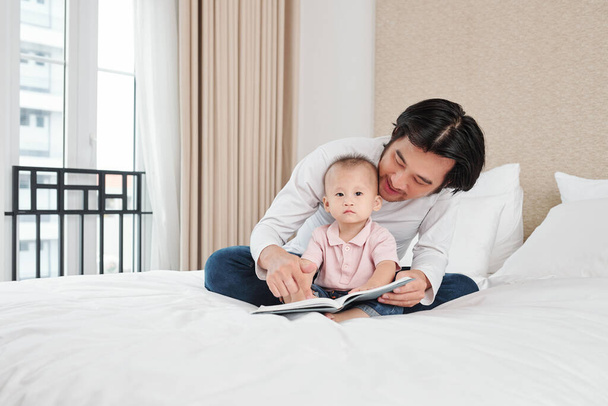 Happy father sitting on bed and reading book for his adorable little son - Photo, Image