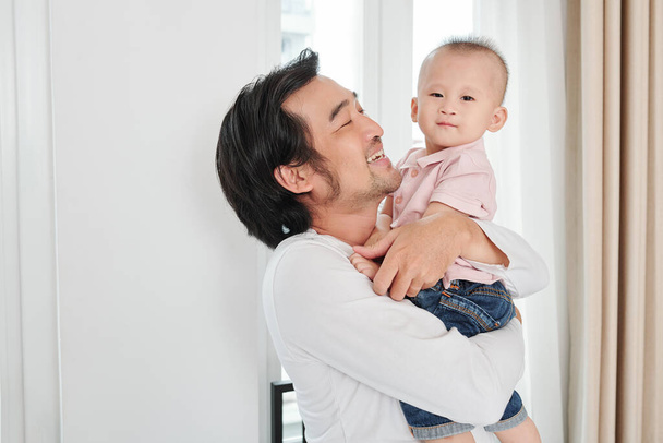 Glücklicher Vietnamese, der die Zeit mit seinem kleinen Sohn genießt. Er trägt ihn und schaut mit Liebe und Zärtlichkeit - Foto, Bild