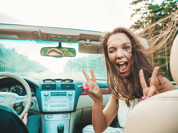 Portrait of two young beautiful and smiling hipster girls in convertible car. Sexy carefree women driving cabriolet. Positive models riding and having fun in sunglasses. Shows peace sign - Photo, image