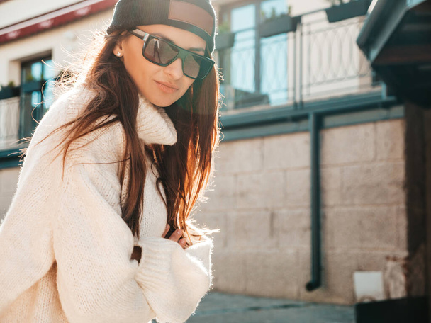 Portrait of beautiful smiling model. Female dressed in warm hipster white sweater and beanie. Trendy girl posing in the street at sunset. Funny and positive woman having fun outdoors in sunglasses - Photo, image