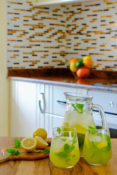 Vintage jug with two glasses of refreshing non alcoholic mojito lemonade drink with lemon slices, mint leaves, straw & ice on wooden kitchen table. Close up, copy space, background - Photo, image