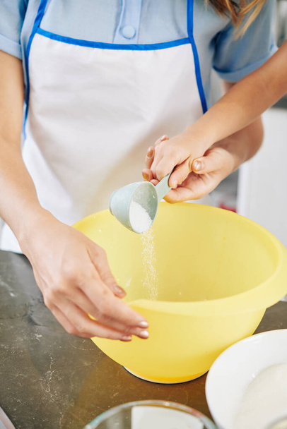 Mère aidant la petite fille à mettre une demi-tasse de sucre dans un grand bol en plastique lors de la fabrication de la pâte à crêpes - Photo, image