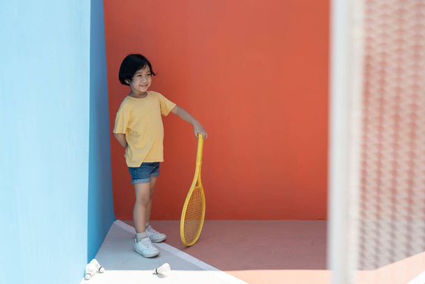 Netter kleiner Junge in gelbem Hemd mit Tennisschläger vor buntem blauem und rotem Hintergrund. Federhähne auf dem Boden. Gelber Tennisschläger in der Hand. - Foto, Bild