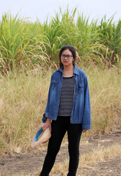 Женщина-фермер, стоящая с соломенной шляпой в руке и в джинсовой рубашке с длинным рукавом на фоне фермы сахарного тростника. - Фото, изображение