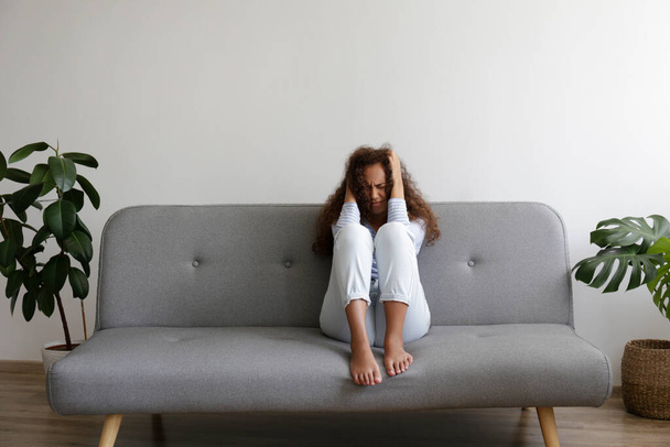 Portrait of young beautiful black woman with depressed facial expression sitting on the couch touching her temples. Female in physical and emotional pain. Sad girl in her room. Background, copy space. - Foto, Bild
