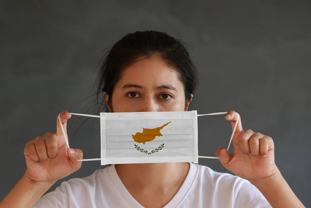 Een vrouw in wit shirt met Cyprus vlag op hygiënisch masker in haar hand en tilde de voorkant op donkergrijze achtergrond. Kleine deeltjes of virus corona of Covid 19 bescherming. Begrip "ziektebestrijding". - Foto, afbeelding