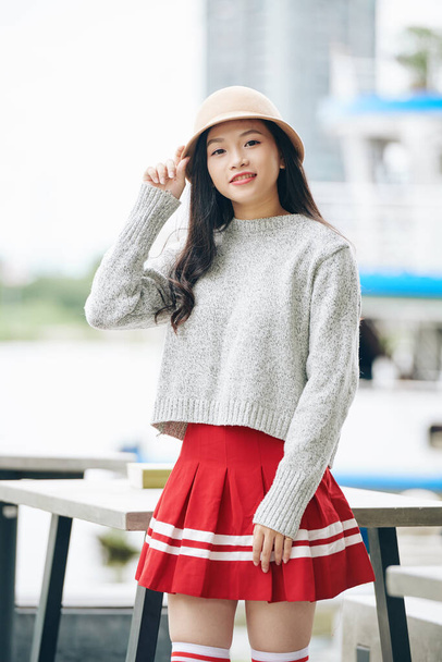 Retrato de sorridente asiático estudante em mini-saia, suéter e chapéu posando ao ar livre
 - Foto, Imagem