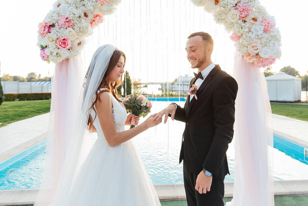 O belo par luxuoso, o noivo em um terno preto e a noiva em um vestido branco de casamento, trocaram anéis em uma cerimônia de saída com um arco, casamento na queda
 - Foto, Imagem