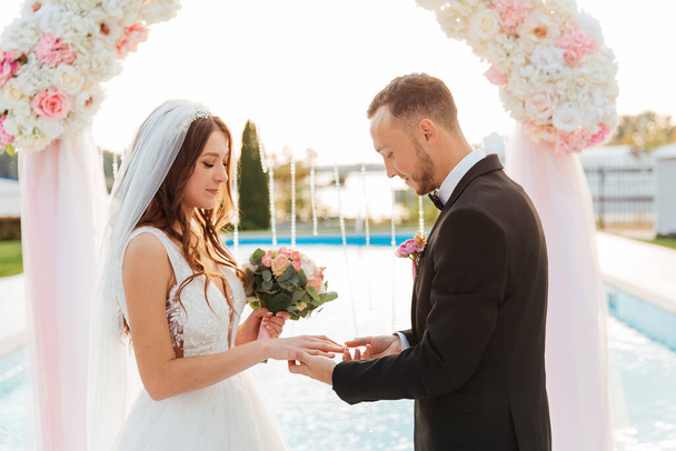 Luxurious beautiful couple, the groom in a black suit and the bride in a wedding white dress, exchanged rings at an exit ceremony with an arch, wedding in the fall - Фото, изображение