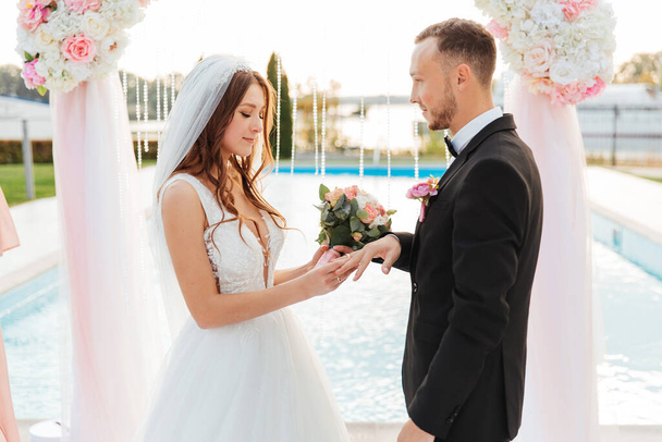 Luxusní krásný pár, ženich v černém obleku a nevěsta ve svatebních bílých šatech, vyměněné prsteny na výstupním ceremoniálu s obloukem, svatba na podzim - Fotografie, Obrázek