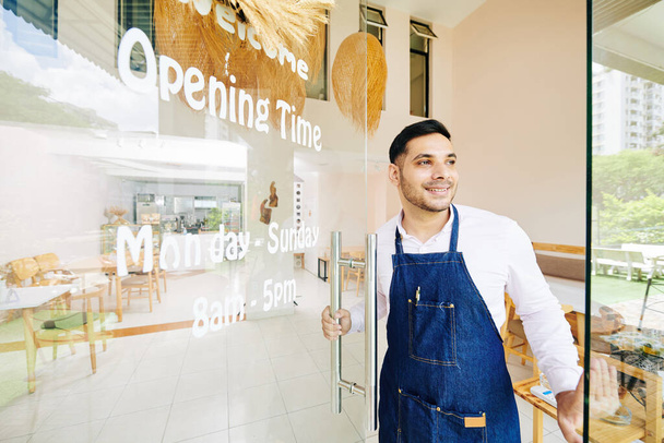 Knappe glimlachende kleine ondernemer openen cafe deur en gastvrije klanten - Foto, afbeelding