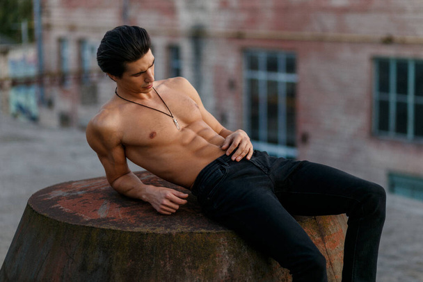 Сексуально привлекательный мужчина с обнаженным туловищем в джинсах, лежит, смотрит вниз, позирует на крыше. - Фото, изображение