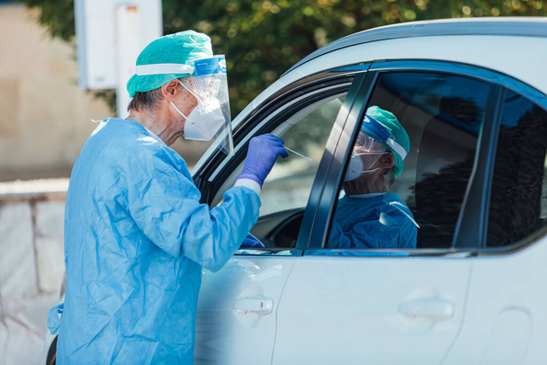 Personnel médical portant un EPI, effectuant une PCR avec un écouvillon à la main, sur un patient à l'intérieur de sa voiture pour détecter s'il est infecté par COVID-19 - Photo, image