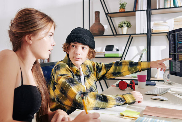 Teenage chlapec v kostkované košili a klobouku ukazuje na obrazovce počítače s programovacím kódem, když dělá domácí úkoly se svou sestrou nebo přítelem doma - Fotografie, Obrázek