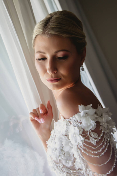 Morning of bride. Slender female in wedding dress at window. Bride look on light. Beautiful wedding morning. Diagonal image. Amazing girl. Woman show emotions. Blonde with blue eyes - Foto, immagini