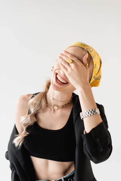 fashionable woman in yellow headscarf and golden accessories covering face with hand and laughing isolated on white - Valokuva, kuva